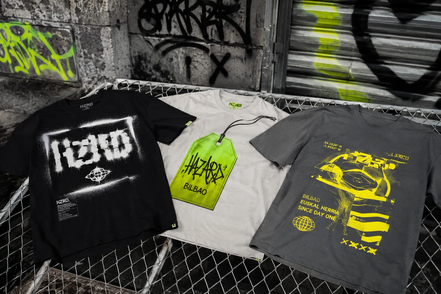 Tres camisetas graficos HZRD colgadas en una valla de tela metalica